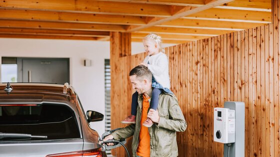 Mädchen sitzt auf den Schultern ihres Vaters, der sein E-Auto auflädt